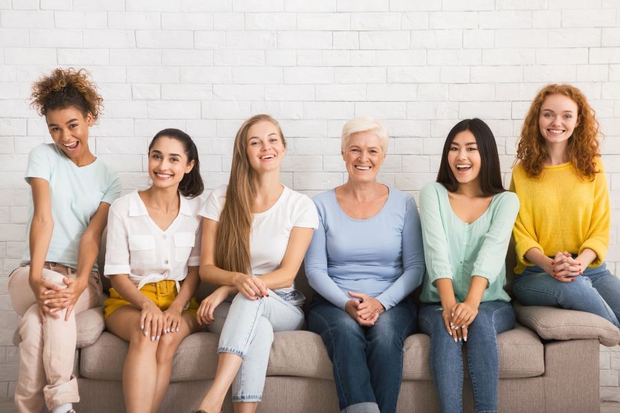 Several happy women of all ages and demographics sitting on a couch and laughing.